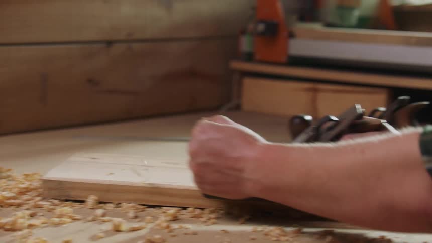 A closeup view focuses on a carpenter wearing a plaid shirt using a metal hand plane to smoothly shave the surface of a piece of light wood on a workbench creating piles of curled wood shavings around the work area