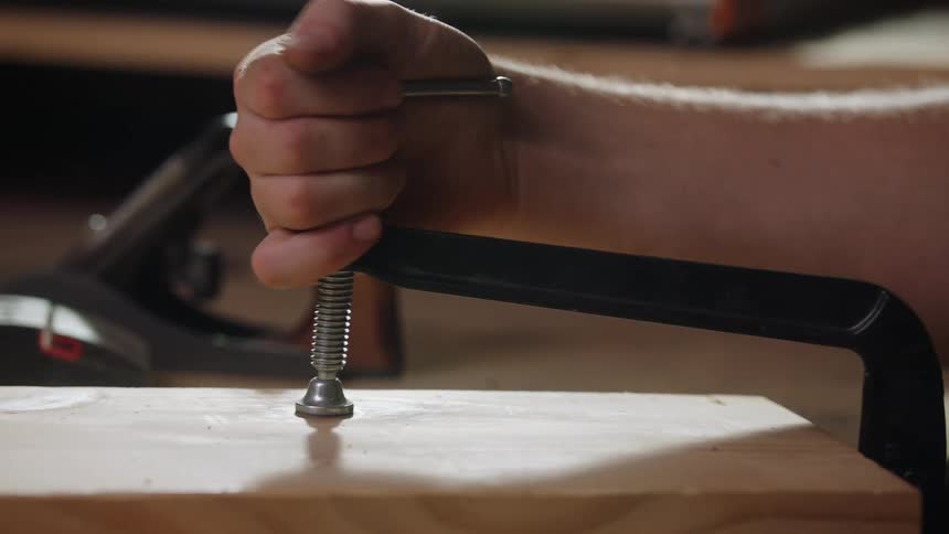 A closeup shot focuses on a black cclamp securing a piece of lightcolored wood to a workbench as a persons hand turns the metal screw mechanism to tighten the grip