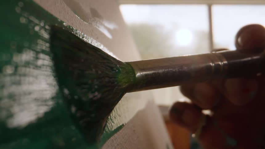 A person is captured in a closeup shot as her hand steadily guides a paintbrush heavily loaded with rich teal paint across a textured lightcolored surface while bright light streams from a window in the background