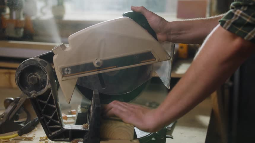 A man wearing a plaid shirt uses both hands to pull down the head of an electric miter saw cutting through a piece of lumber on a workbench covered in sawdust in a brightly lit workshop
