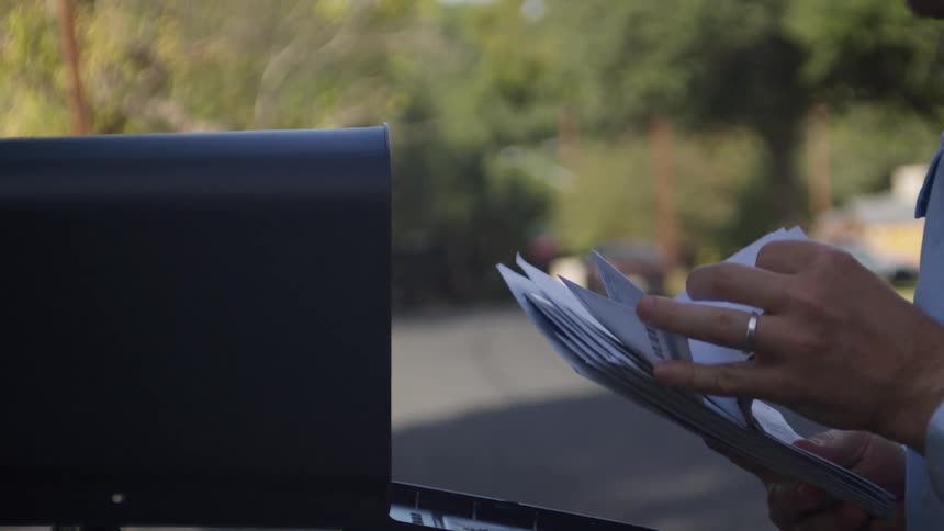 A mail carrier approaches a black roadside mailbox opens the door to place several letters inside and then closes it before continuing his delivery route