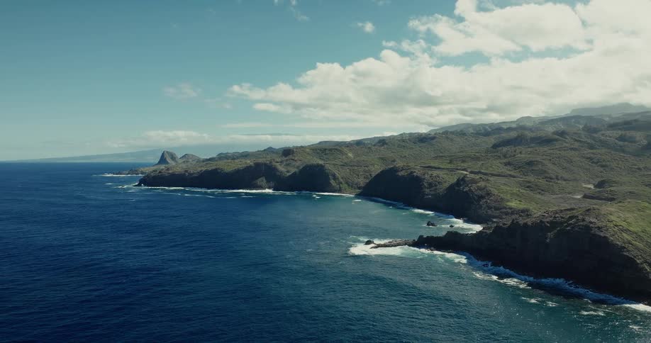 The view slowly pulls back over a deep blue ocean revealing a vast and rugged green coastline where white waves crash against dramatic rocky cliffs