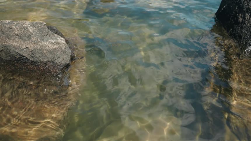 Clear water ripples gently around dark rocks as sunlight creates shimmering patterns on the shallow riverbed below
