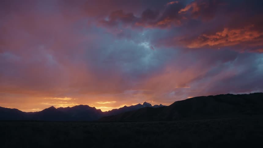 A mountain range stretches across the horizon with an orange cloudy sky glowing above it at sunset