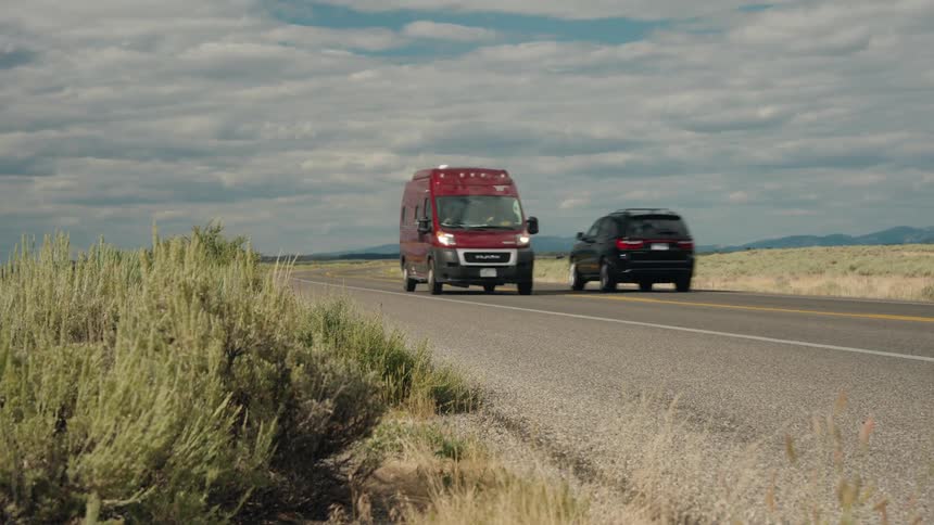 A red van drives down a highway through a dry grassy landscape under a cloudy sky