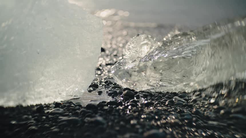 A close view of meltwater streaming off a clear iceberg fragment onto dark shining pebbles between two larger pieces of glistening ice