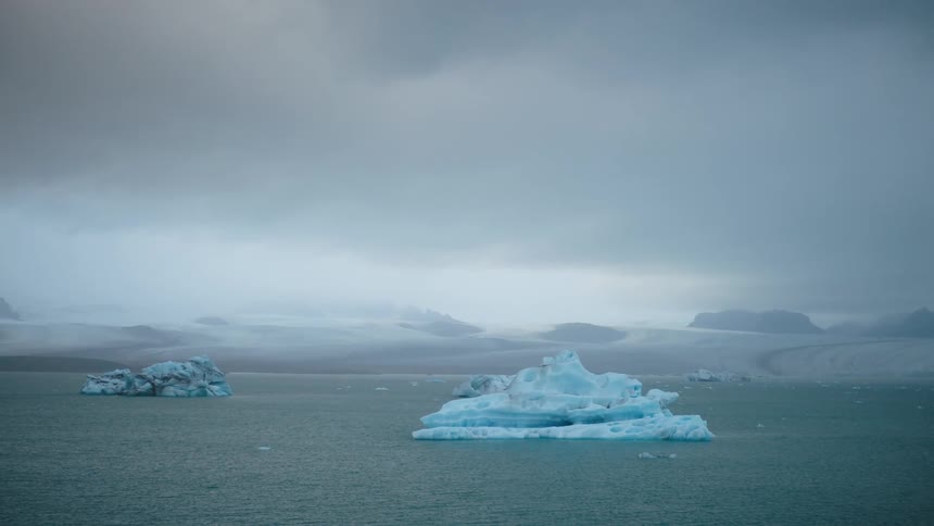 A blue icebergs drift through calm misty arctic waters under overcast skies