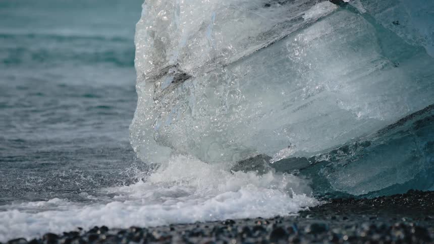 A close view of a massive chunk of blue ice on dark wet sand as the ocean waves crash and foam splashes around its base