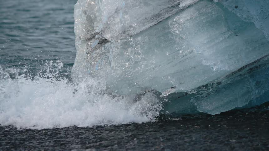 A close view of a massive chunk of blue ice on dark wet sand as the ocean waves crash and foam splashes around its base