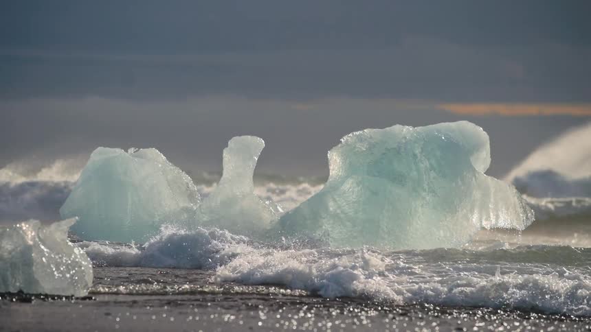 Ocean waves hitting ice chunks on rocky shore at dusk