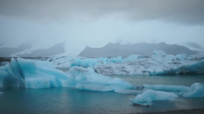 Blue and white icebergs floating in cold water with mountains in background under cloudy sky