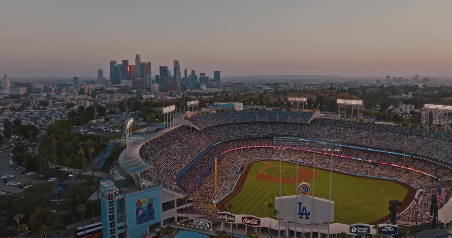 A wide highangle aerial shot captures a baseball stadium during a game with its bright green field and crowds illuminated against the hazy dusk sky and the distant skyline of acity