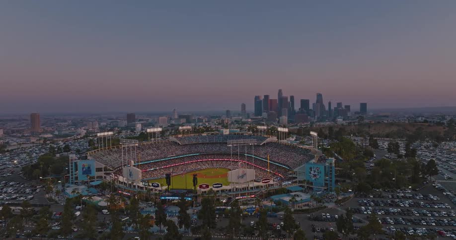Aerial view of a packed baseball stadium glowing at dusk with a city skyline behind it