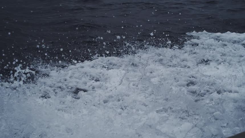 The wake of a boat churns the dark ocean water sending a chaotic spray of white foam and droplets into the air in slow motion
