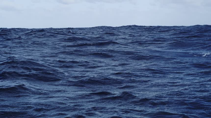 A static shot captures the rough turbulent surface of the deep blue ocean where numerous waves and ripples move constantly beneath a pale cloudy sky that meets the water at the horizon