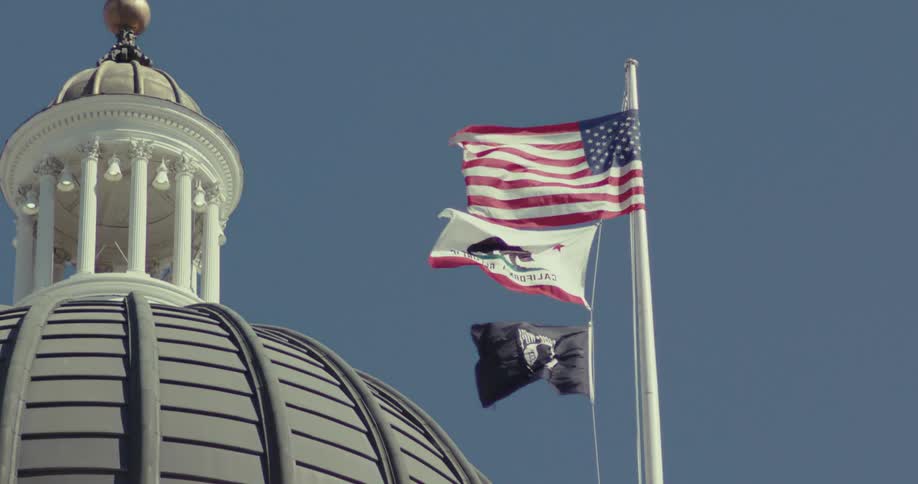 An american flag flies on a pole beside two other flags near a tower at midday