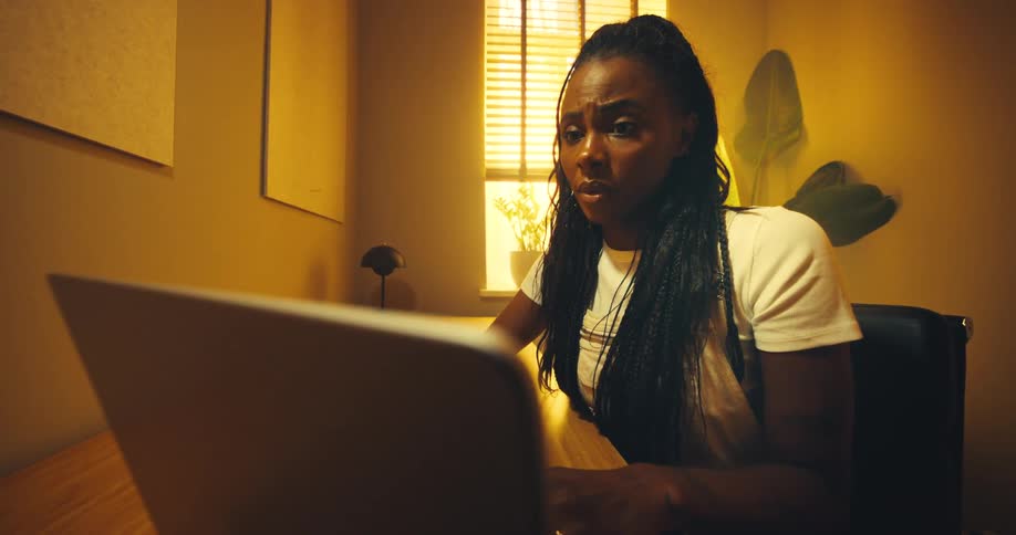 A young woman with braids sits at a desk working on her laptop in a room illuminated by warm golden light