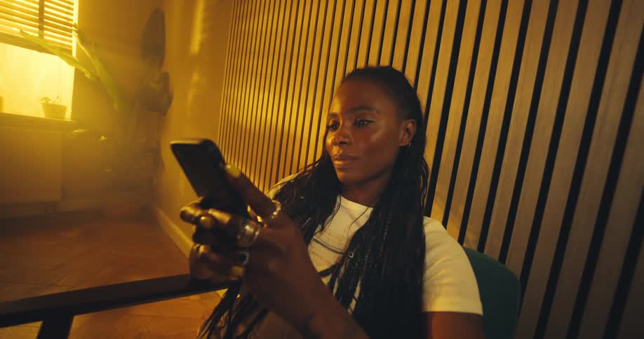 A woman with long braids wearing a white shirt and several rings sits in a chair against a background of warm vertically slatted wood and brightly backlit window light as she focuses intently on her smartphone