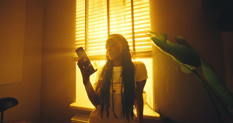 An overhead shot transitions to a medium closeup of a young woman with long dark braids standing in a brightly sunlit room with sloped walls looking down at the mobile phone she holds in her hands and smiling