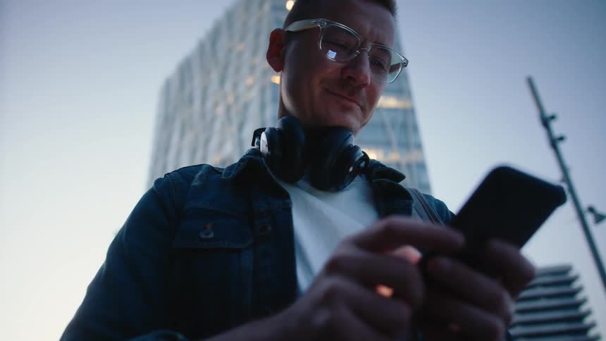 A man with glasses and headphones smiles as he uses his smartphone in front of a modern city skyscraper