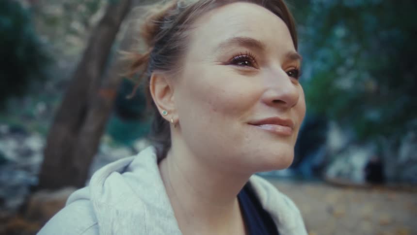 A closeup shot features a smiling woman with her hair pulled back looking upwards and offcamera in a natural outdoor environment the background is softly focused showing lush green foliage and the faint presence of a waterfall or rushing water