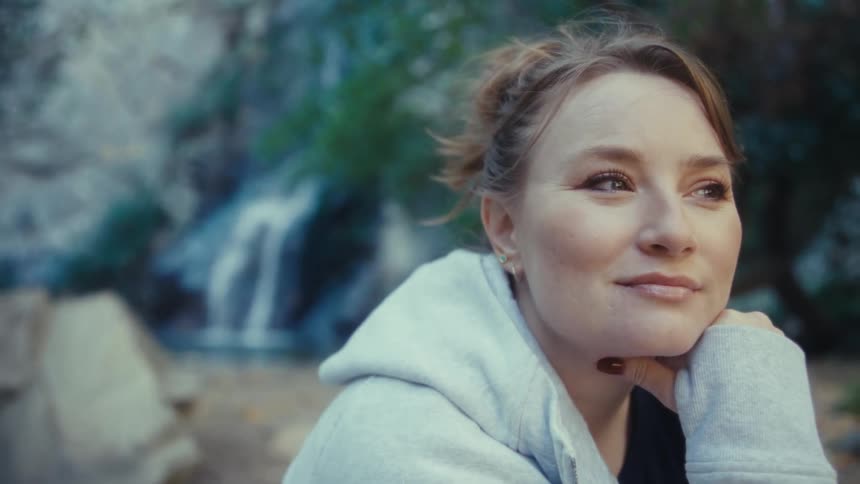 The video opens with a medium shot of a woman seated on a rock in a wooded area leaning on her hand and looking thoughtfully into the distancethe camera slowly zooms in on her face as she closes her eyes for a moment then opens them with a serene exp