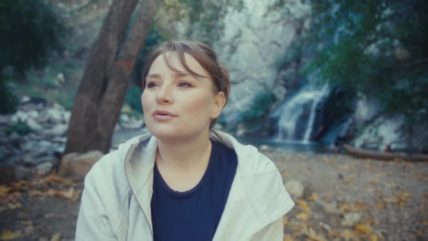 A woman sits serenely on the forest floor momentarily closing her eyes to take a deep breath before opening them with a slight smile as she enjoys the natural environment with a tiered waterfall visible in the densely wooded background