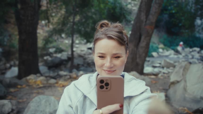 Woman holds her phone as she stands among trees and boulders on a mountain taking a picture of an orange leaf in her hand