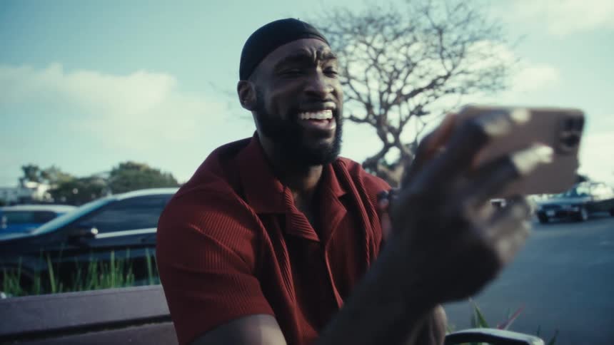 A man is sitting on an outside bench looking at his cell phone and he is happy and then celebrates by pointing upwards