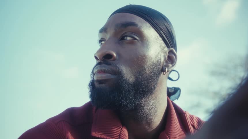 A man wearing a durag and a red shirt is shown outdoors initially looking intently to the side he then turns to the camera smiles and holds up his smartphone with a tan case suggesting he is taking a selfie or recording a video