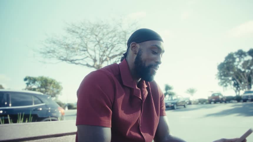 A smiling black man wearing a red shirt and black cap sits on a park bench looking down and interacting with his mobile phone on a sunny day