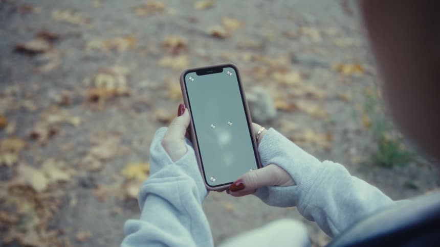 This video shows a closeup of a person holding a smartphone outdoors the person wearing a lightcolored sweatshirt or jacket scrolls or taps on the phones screen which is blankgreenscreened for mockup purposes with fallen autumn leaves visible i