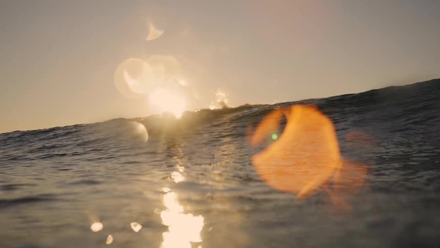A surfer paddles over the crest of a wave in the golden light of sunset viewed from the water level