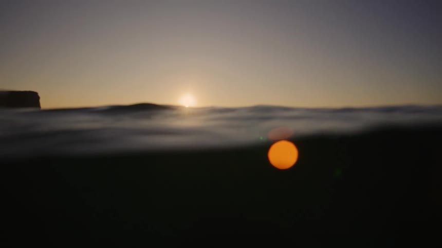 Distant surfers float on the swelling ocean surface silhouetted against a bright golden sunset