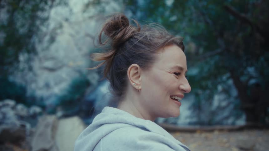 A smiling woman waves and talks on a video call in a forest turning to show the scenery behind her