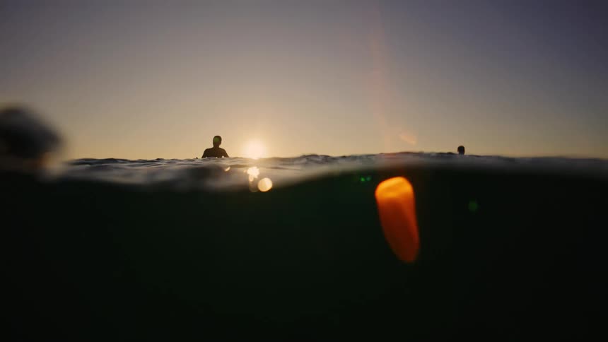 Silhouettes of two surfers sitting on their boards in the ocean framed by the setting or rising sun directly in the center which casts a bright golden reflection and flare on the water surface and a dark silhouette of a cliff on the left