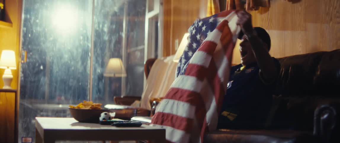 An elderly man is sitting on a sofa and then begins to celebrate and wave an american flag in front of him