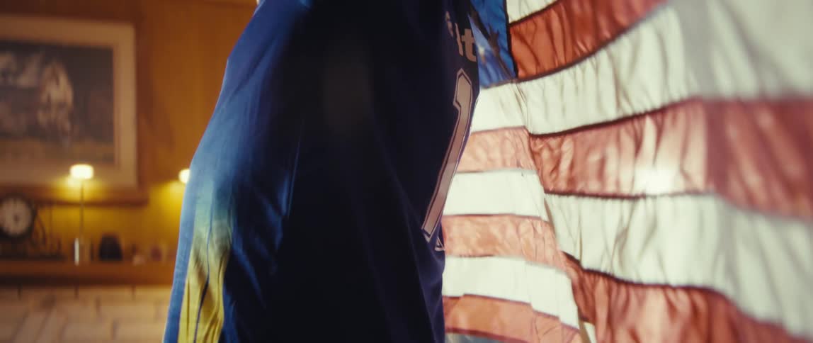 An elderly man wearing a baseball cap and draped in an american flag turns away from the camera