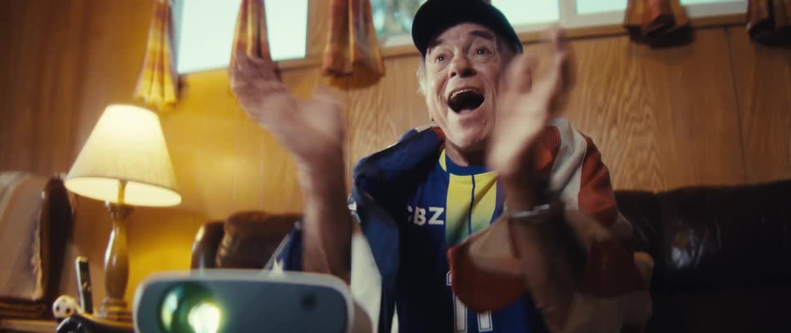 An older man wearing a blue sports jersey a cap and an american flag draped around his shoulders enthusiastically shouting and raising his fists while watching a projected game in a dimly lit retrostyle living room