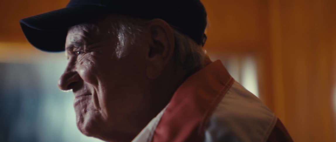 Elderly man in black cap and redgray jacket laughing in warmly lit indoor setting
