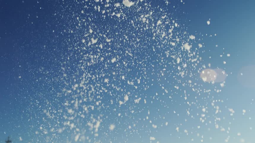 A shower of bright white snow or fine particles is dramatically tossed high into the air against a clear deep blue skythe particles catch the sunlight creating a sparkling effect and lens flare as they drift and fall against the bright unclouded bac