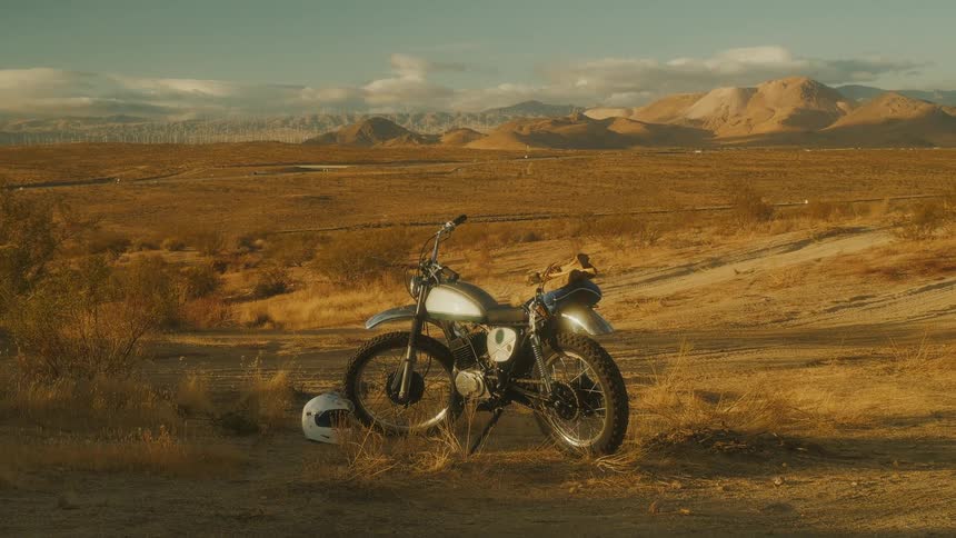 A motorcycle stands parked in a vast sun drenched desert landscape with a helmet resting on the ground beside it