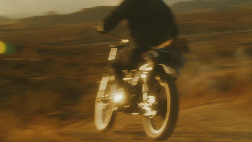 The video shows a motorcyclist riding a vintagestyle bike rapidly across a dusty offroad desert landscape the camera starts close on the wheel and then pans out to reveal the rider who is wearing a black outfit and a white helmet kicking up a trail