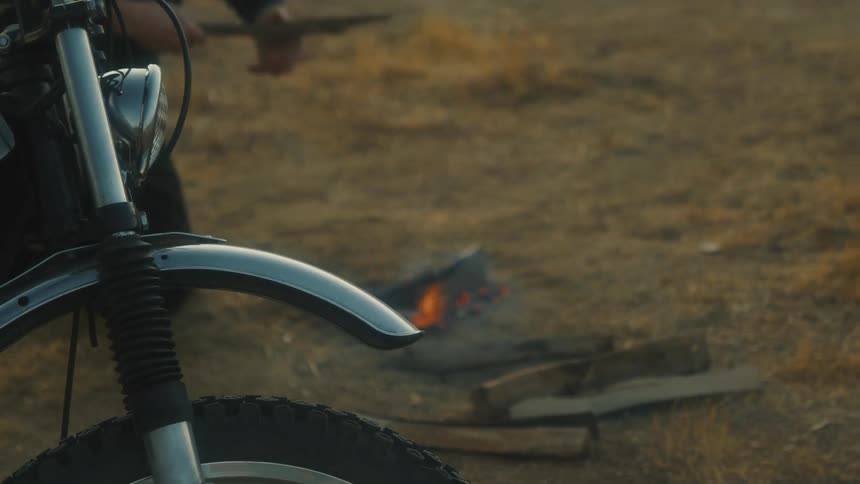The front of a motorcycle is visible while a person tends to a small campfire in a grassy field
