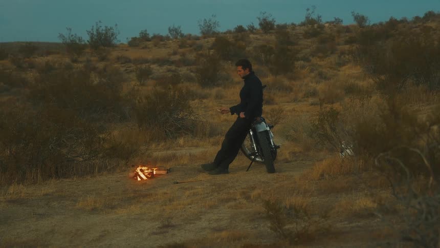 The video shows a man standing next to his motorcycle in a dry scrubland desert environment possibly at twilight he is facing a small burning campfire on the ground next to the bikes rear wheel