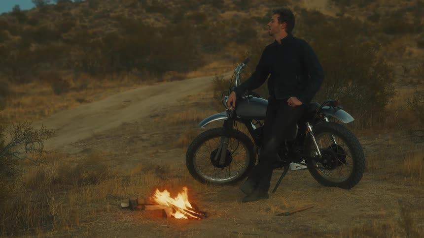 The man is smoking while leaning on his motorcycle by the campfire