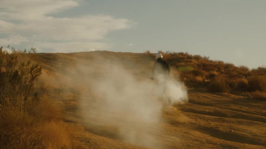 A man in a jacket and white helmet rides his bike up a hill in the valley at midday with smoke trailing behind