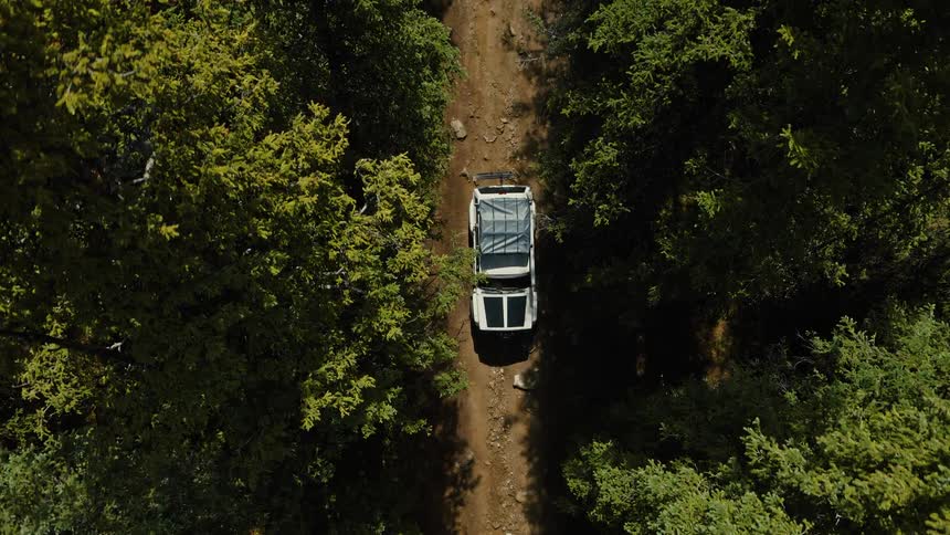 The video is an overhead drone shot showing a white offroad vehicle possibly a classic 4x4 with a roof rack driving slowly along a narrow unpaved dirt road the road is deeply carved between dense walls of tall green trees suggesting a remote mountai