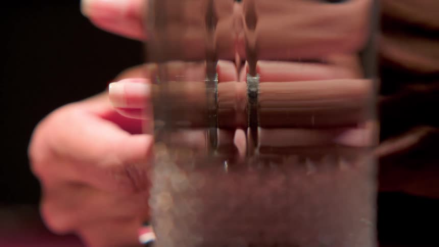 A woman wears a diamond ring on her finger with a glass positioned in front of her hands sitting in a bright room contrasted by a dark background