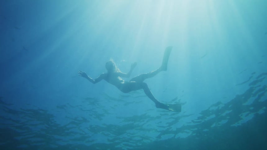 An underwater shot captures a diver wearing fins and a mask descending into the deep blue water silhouetted against the bright surface light before swimming out of the frame